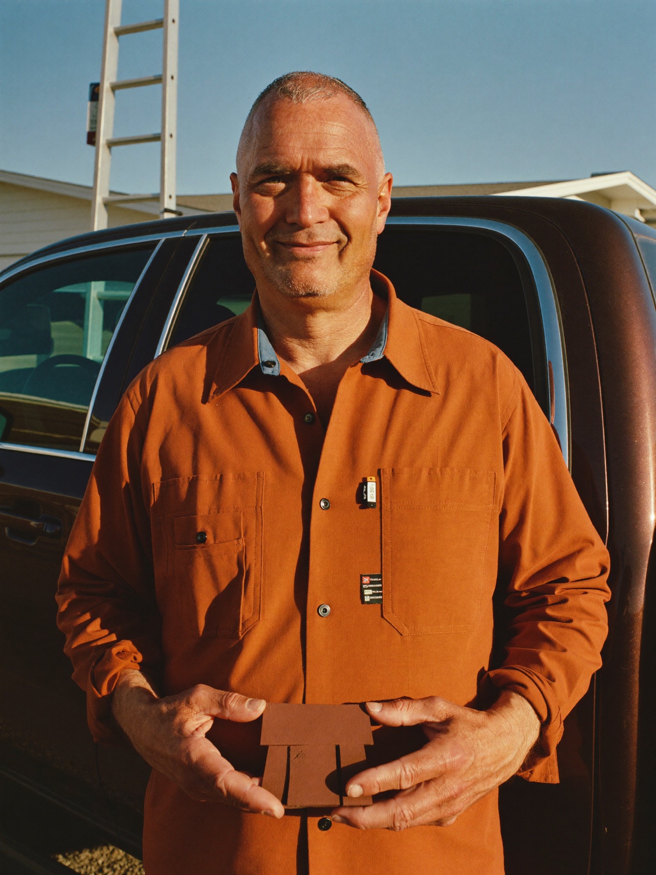 Cole Barrett, the fictional owner of Redbud Roofing, holding a shingle sample by his pickup at golden hour